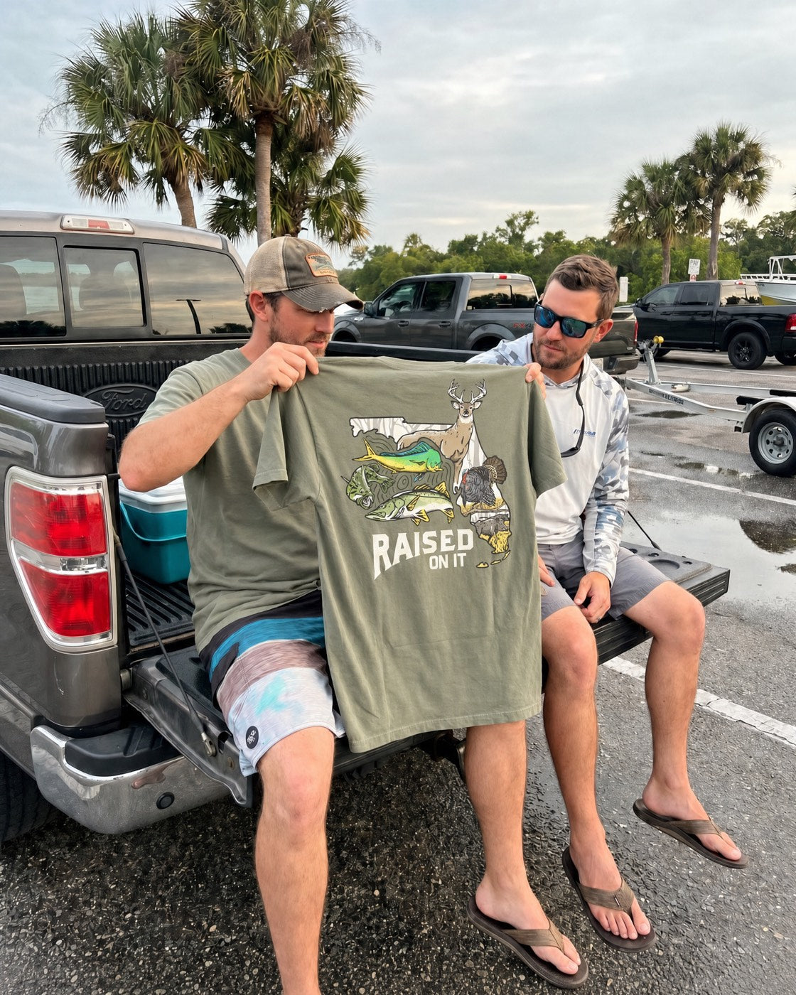 Two friends on a truck tailgate at the boat ramp, one holding up a Florida Sportsman tee