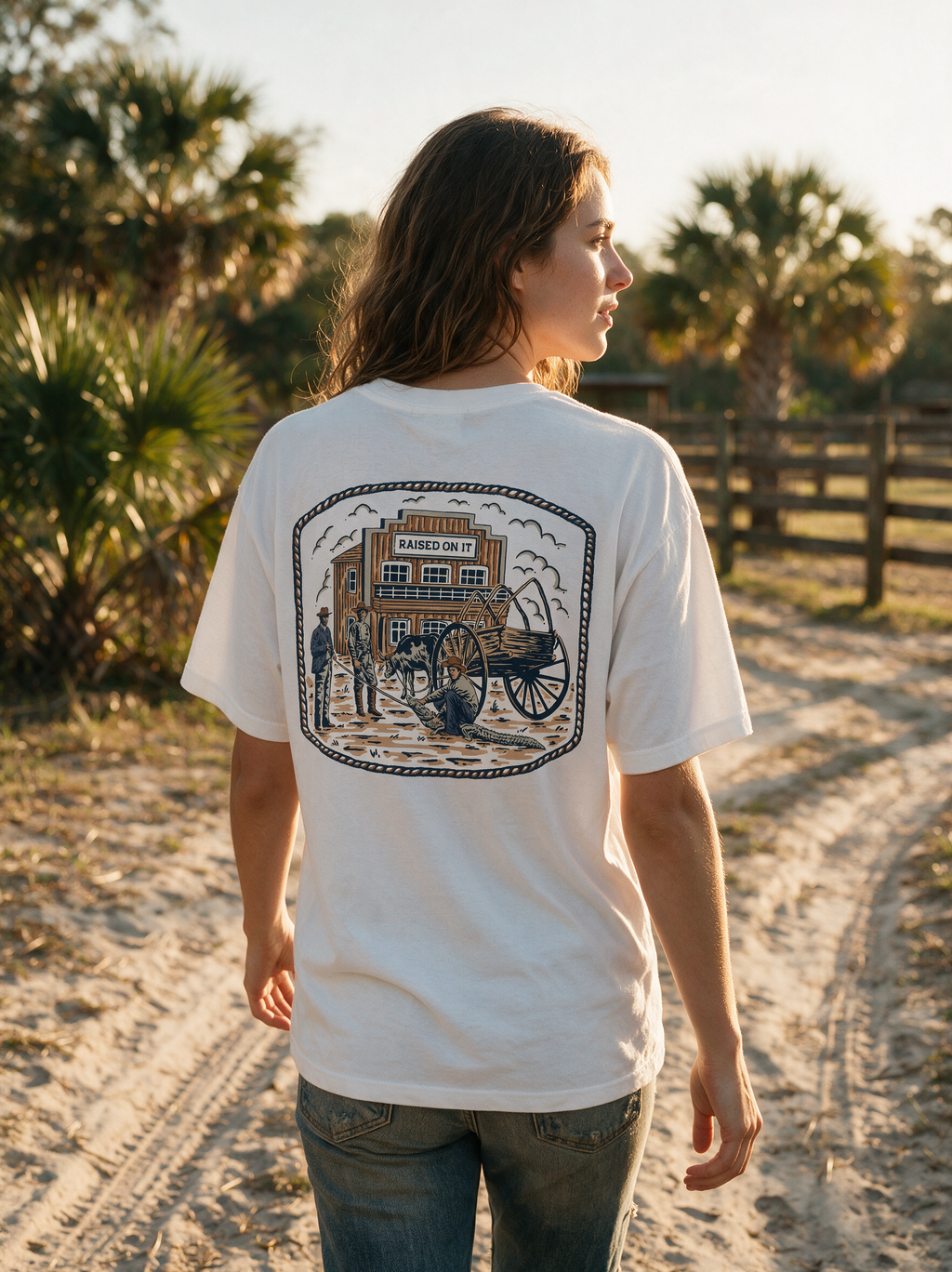 Woman wearing the white Florida Cowboy tee on a sandy ranch road at golden hour, relaxed unisex fit, back print in full view