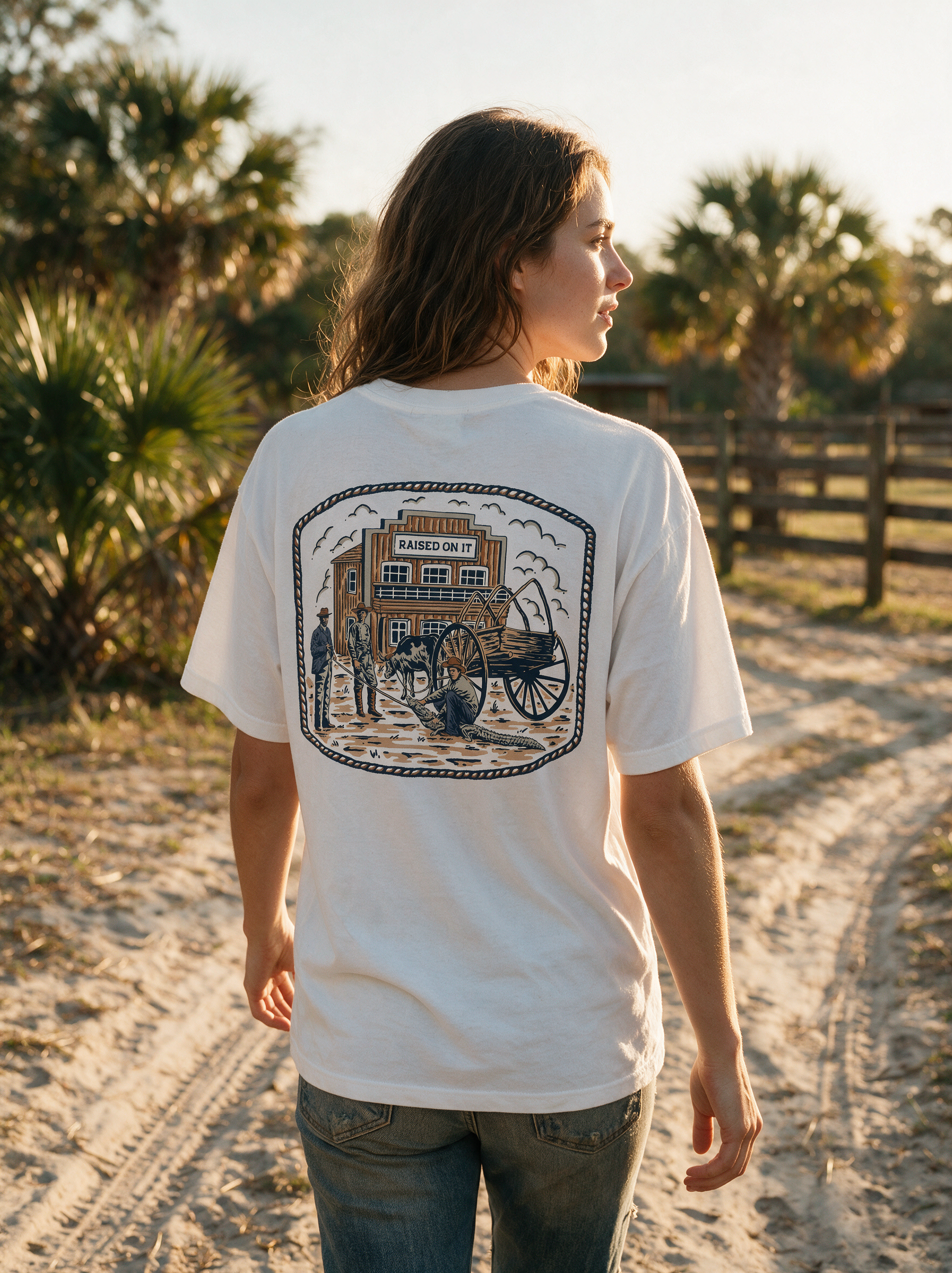 Woman wearing the white Florida Cowboy tee on a sandy ranch road at golden hour, relaxed unisex fit, back print in full view