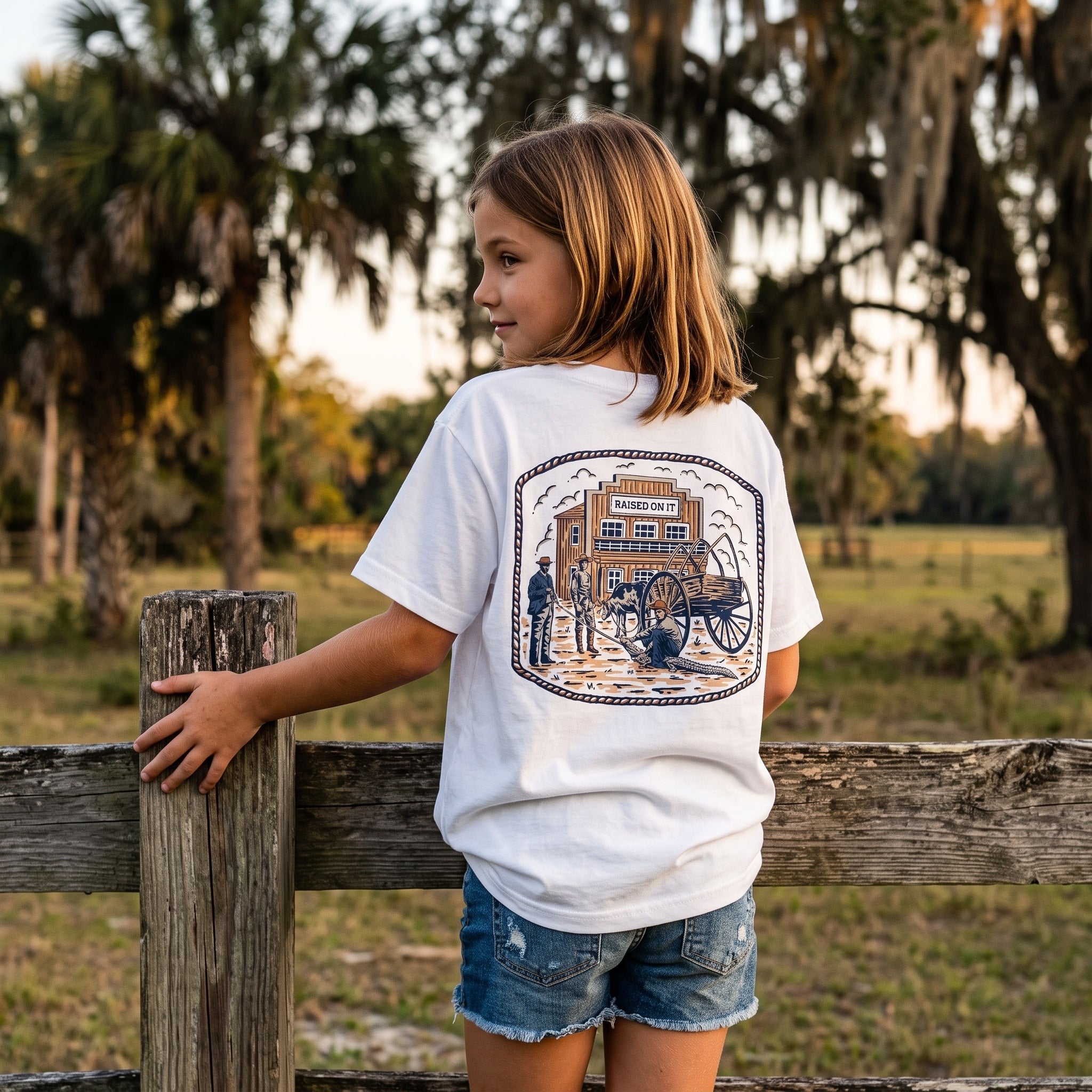Youth Florida Cowboy Tee back print shown on a girl beside a pasture fence under live oaks
