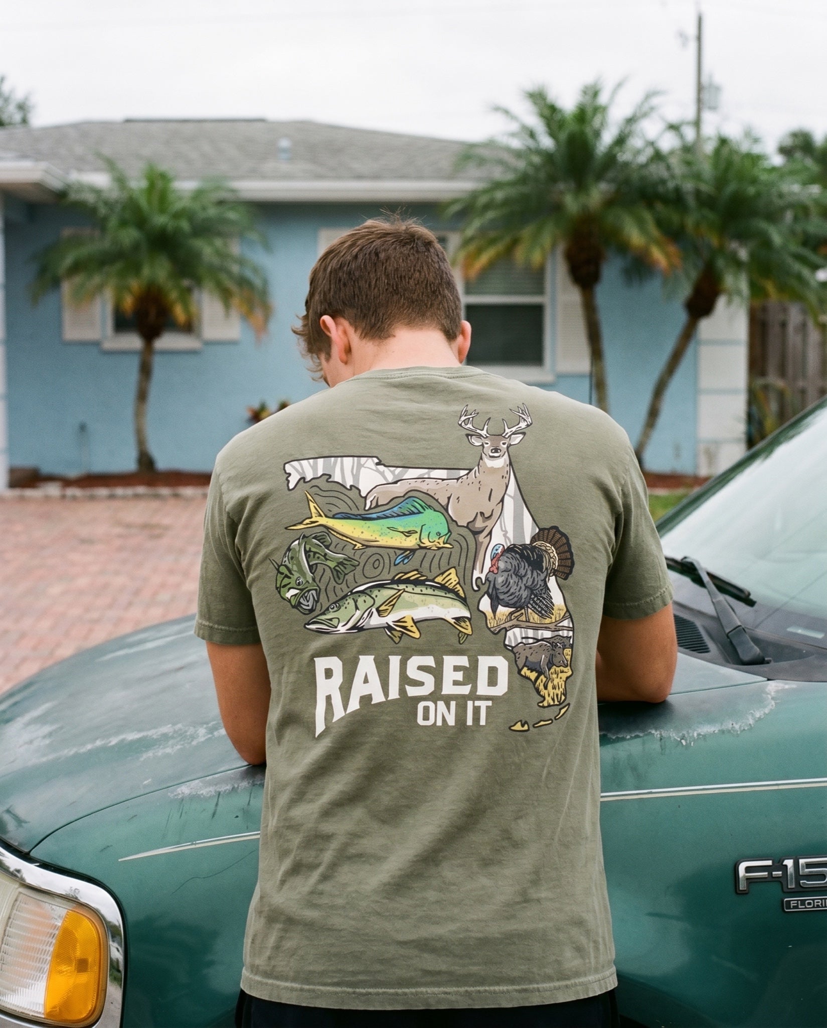 Back view of the olive Florida Sportsman Tee worn leaning against a vintage pickup truck, full wildlife print visible