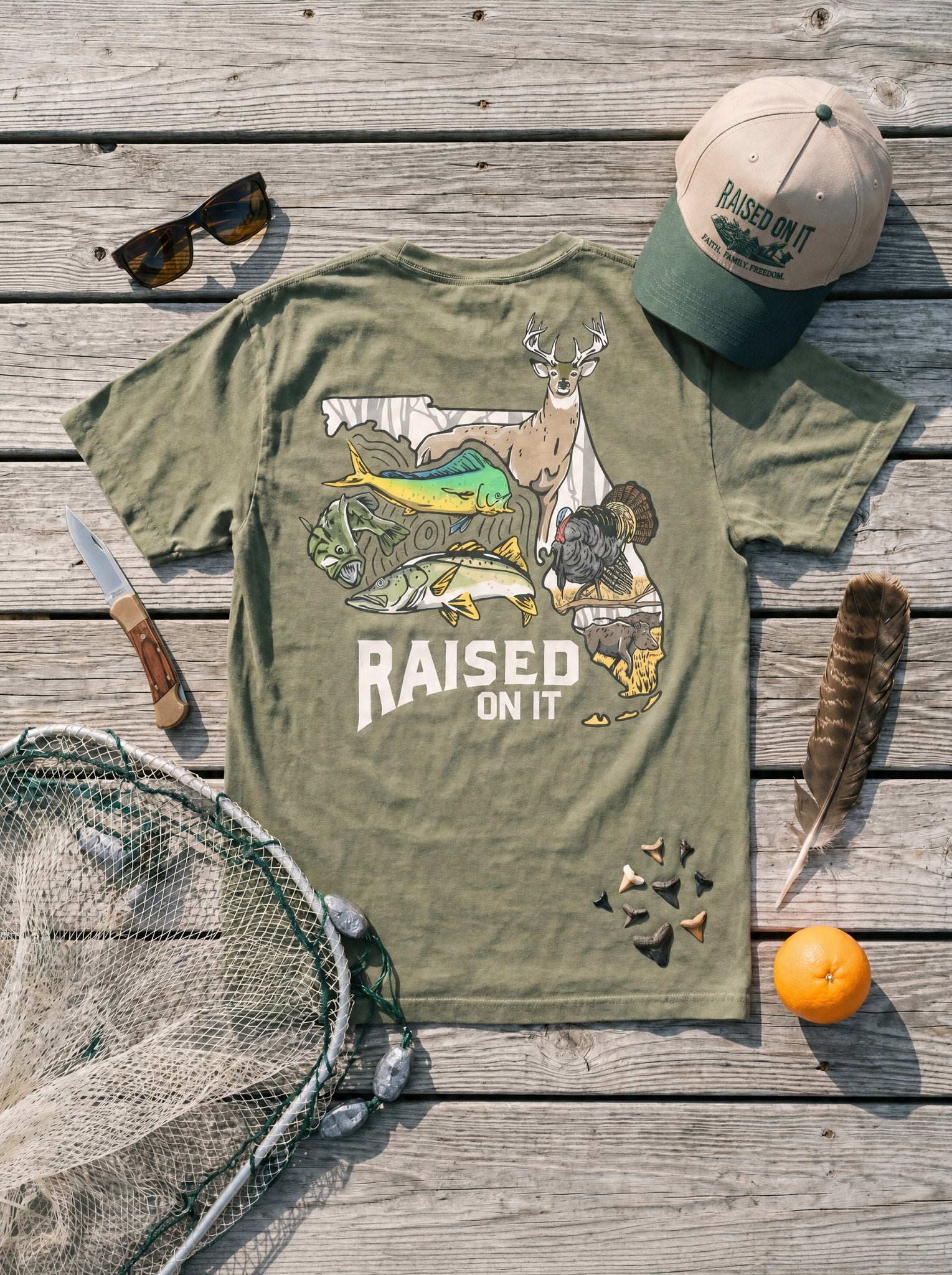 Olive Florida Sportsman Tee flat-lay with cast net, turkey feather, shark teeth and Raised On It hat
