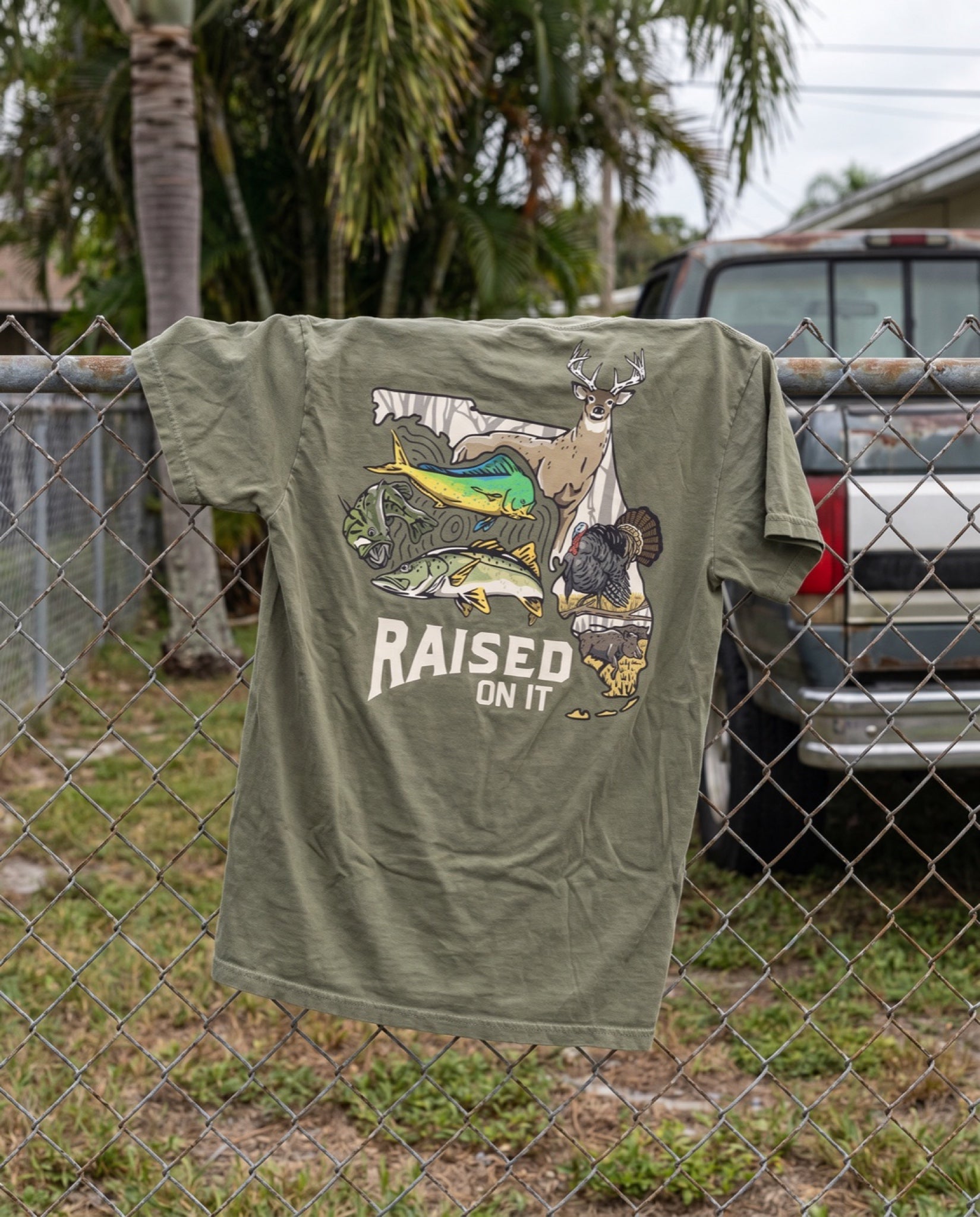 Olive Florida Sportsman Tee draped on a chain-link fence in a Florida backyard, back print visible